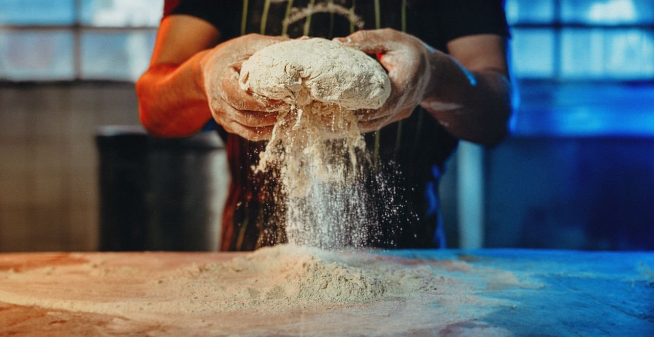 Services A baker works by hand with flour and dough, creating artisanal bread in an industrial kitchen setting.